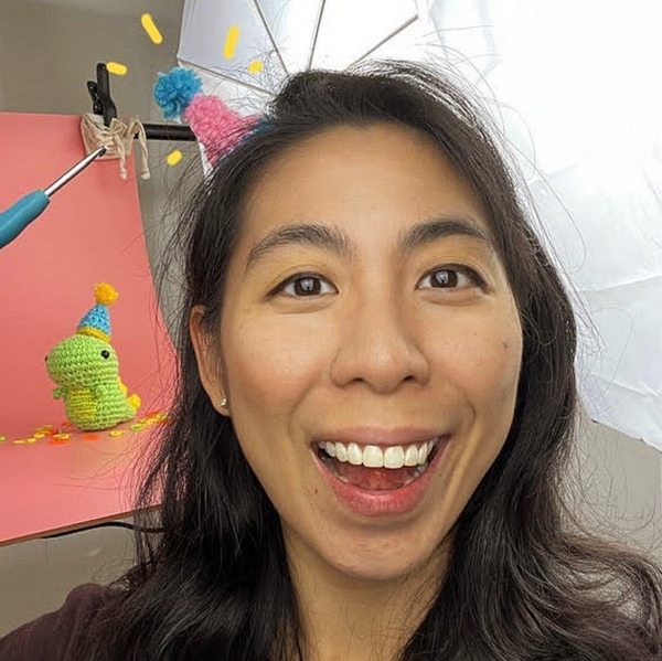 A photo of Chief Woobler at the Woobles, Justine Tiu with an amigurumi dinosaur behind her on a pink backdrop