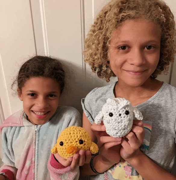Two young sisters standing side-by-side in front of a white door and showing their chick and owl amigurumi plushies